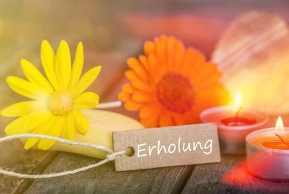 Gelbe und orangefarbene Blüten neben brennenden Teelichtern und einem Etikett mit der Aufschrift Erholung auf einer hölzernen Oberfläche, warmes, beruhigendes Licht.