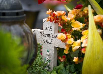Steinernes Kreuz mit der Inschrift 'Ich Vermisse Dich', umrahmt von gelb-orangenen Blüten (Begonien) und grünem Bewuchs, links unscharf eine Grablaterne.