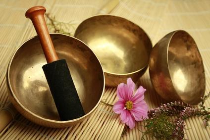 Drei bronzefarbene Klangschalen auf einer Bambusmatte, eine enthält einen Holzschlägel mit schwarzem Filzkopf; daneben eine rosa Blüte und kleine Blütenzweige.