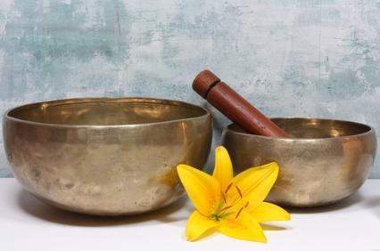 Zwei bronzefarbene Klangschalen unterschiedlicher Größe, bei der kleineren liegt ein brauner Holzklöppel, davor liegt eine gelbe Lilienblüte vor einer leicht strukturierten hellblauen Wand auf weißem Untergrund.