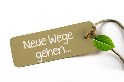 Braunes Etikett mit weißer Schrift 'Neue Wege gehen...', an einer Schnur befestigt und mit einem kleinen grünen Blatt, auf weißem Hintergrund.