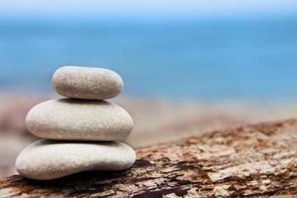 Drei glattgeschliffene helle Kieselsteine aufeinandergestapelt auf einem verwitterten Treibholzstück vor unscharfem blauem Meeres- und Himmelshintergrund.