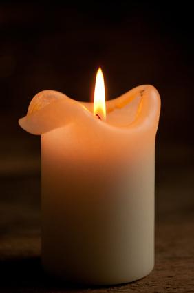 Weiße Stumpenkerze mit geschmolzener, gewellter Oberkante und einer warm leuchtenden Flamme vor dunklem Hintergrund auf einer Holzfläche.